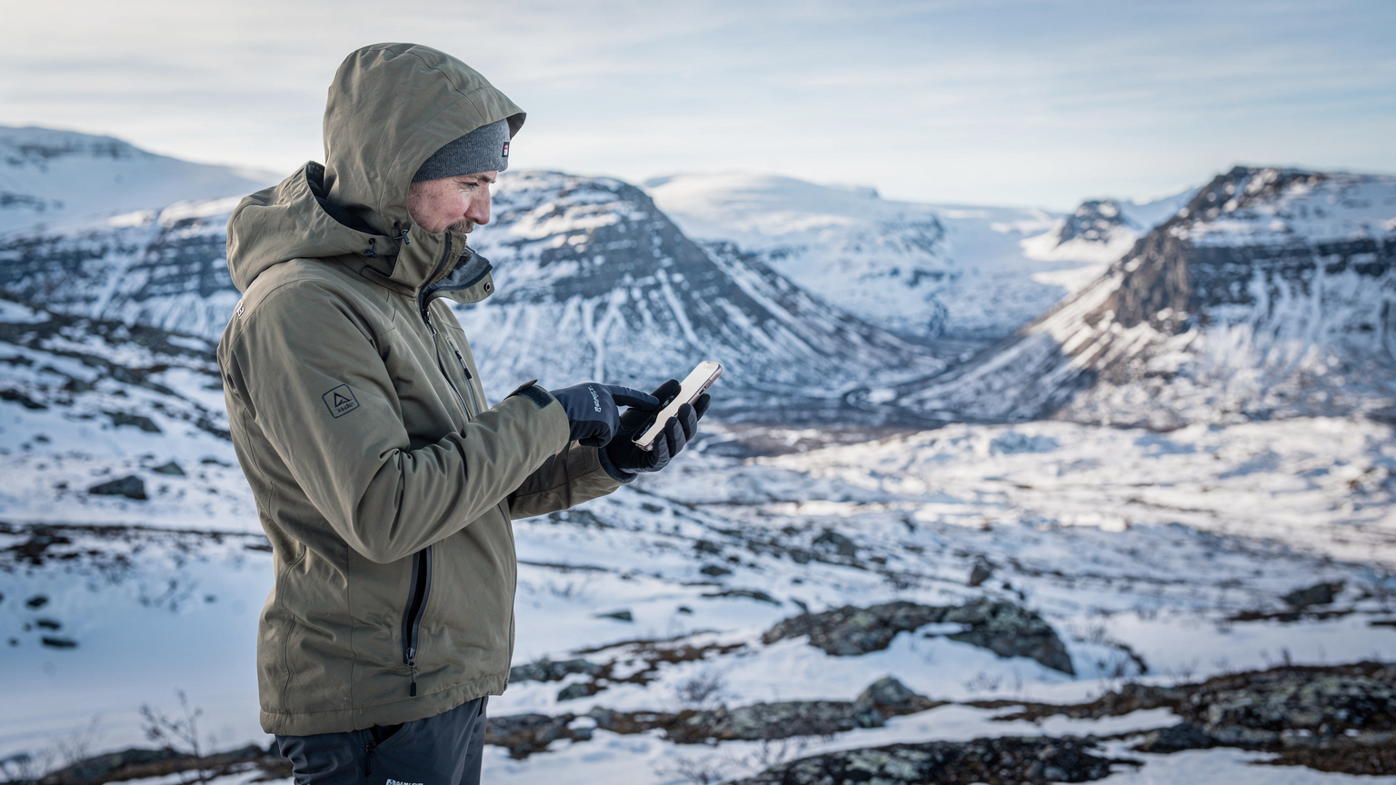 En person i vandringskläder pekar på en digital karta på en smartphone mot en bakgrund av en vidsträckt, snötäckt dal och berg i Sarek nationalpark.