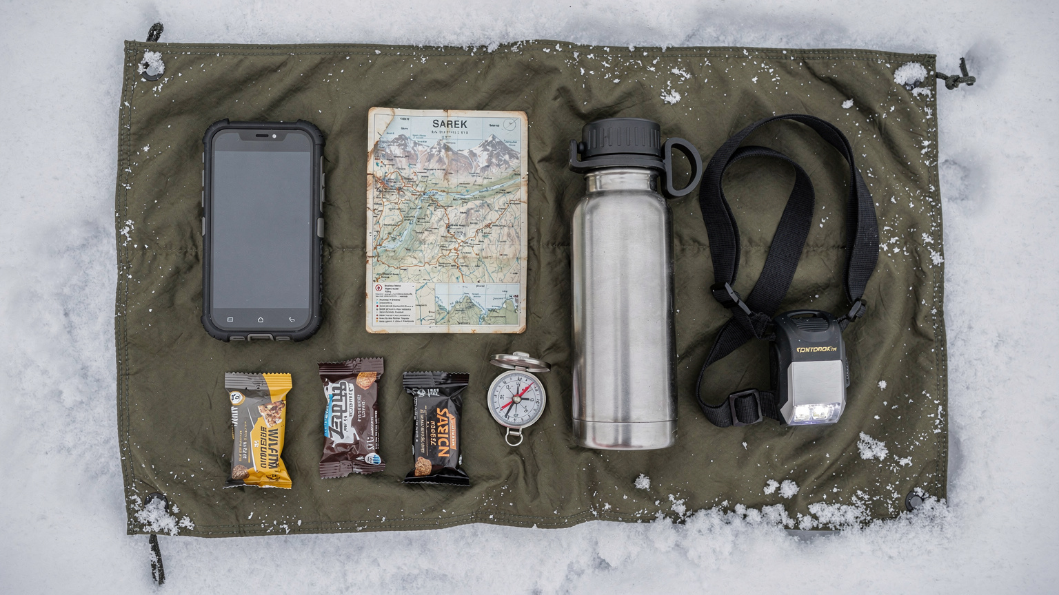 En översiktsbild av expeditionsutrustning utspridd på en snötäckt presenning, inklusive en smartphone, en satellitkarta, en termos, en pannlampa och en kompass.