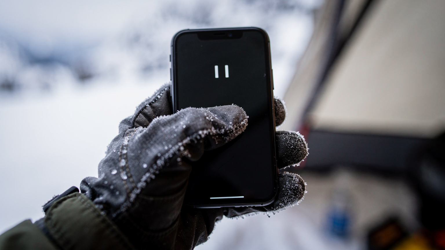 En närbild av en hand i en tjock, frusen handske som håller en smartphone med ett svagt signalfält synligt på skärmen, mot en oskarp, snöig bakgrund.
