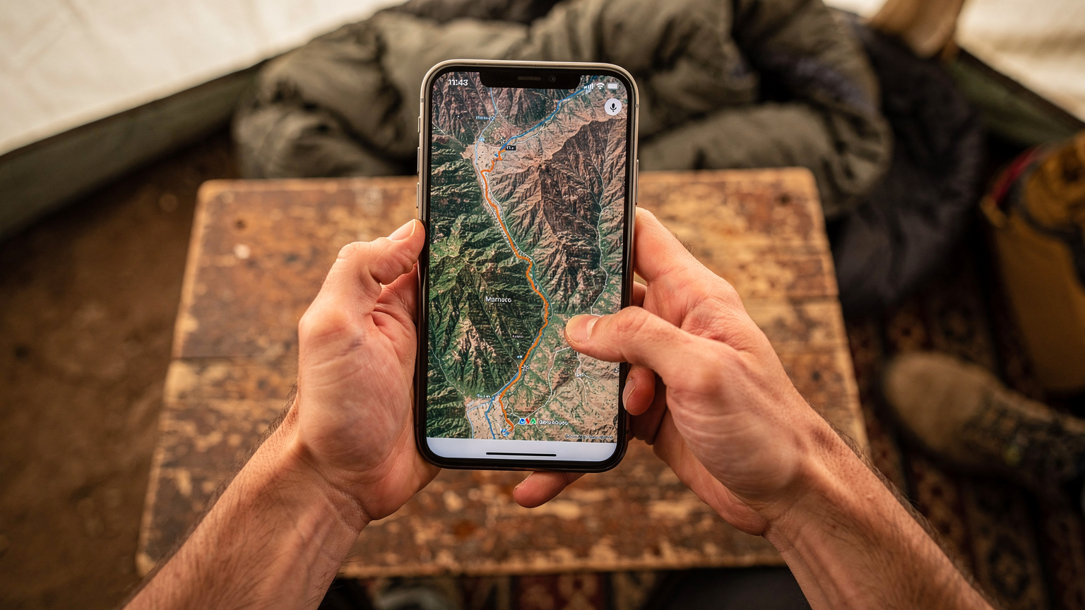 Nærbilde av en reisende sine hender som holder en smarttelefon, med et kart over Marokko og Atlasfjellene åpent på skjermen, mens de peker på en rute med fingeren.