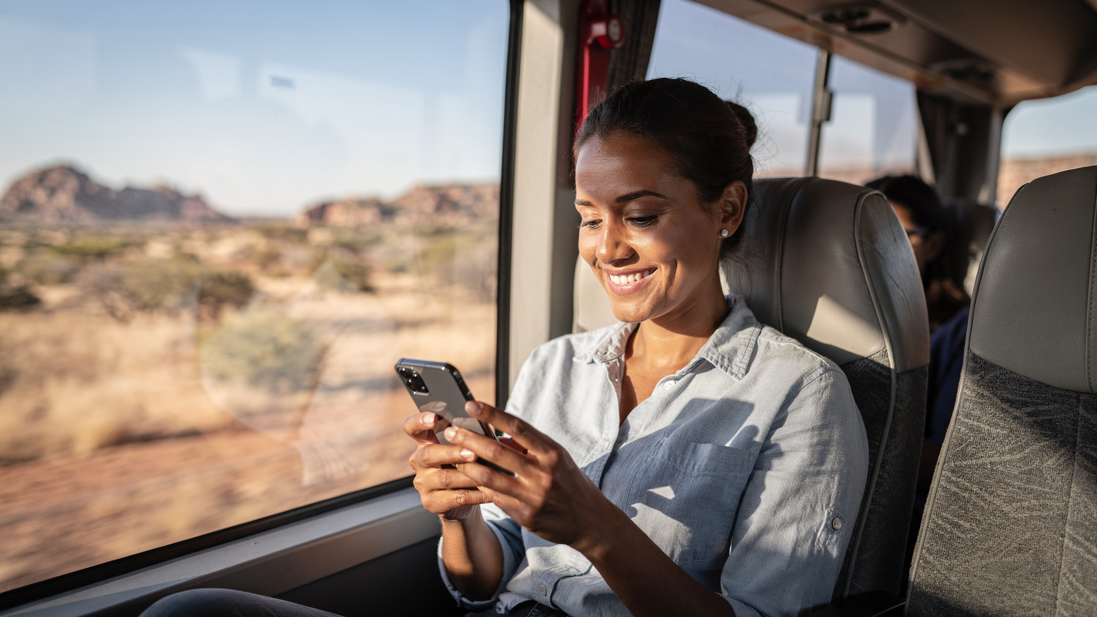 Une femme souriante, les yeux rivés sur son téléphone, naviguant sur une carte, installée confortablement dans un bus interurbain traversant un paysage aride de l'Afrique australe.