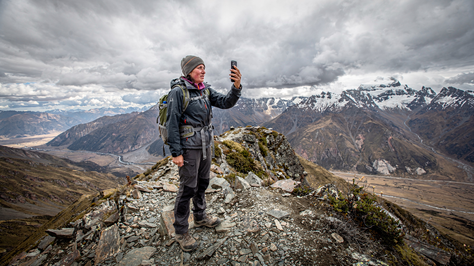 En hiker står på en bjergtop med et let bekymret udtryk, mens hun holder sin telefon op i luften i et forsøg på at fange signal.