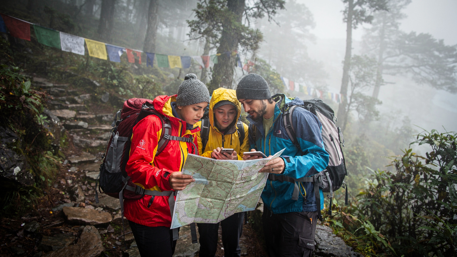 En grupp vandrare i regnkläder rådgör med en karta och en smartphone i ett dimmigt, regnigt bergslandskap i Nepal, vilket illustrerar vikten av navigation under utmanande väderförhållanden.