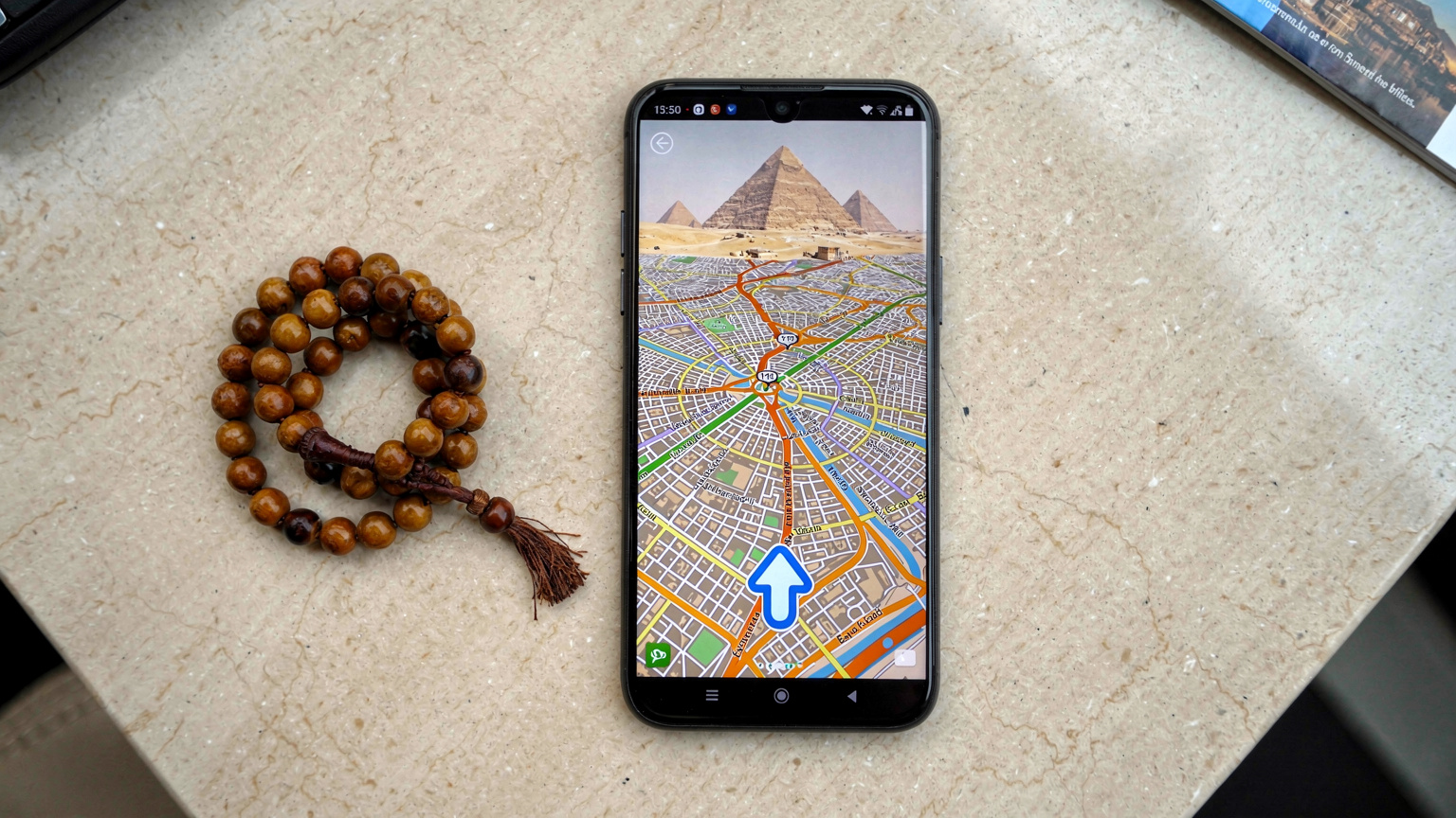 Un navigatore GPS su smartphone mostra una mappa dettagliata delle strade del Cairo, con una freccia blu che indica la posizione corrente e le Piramidi in lontananza.