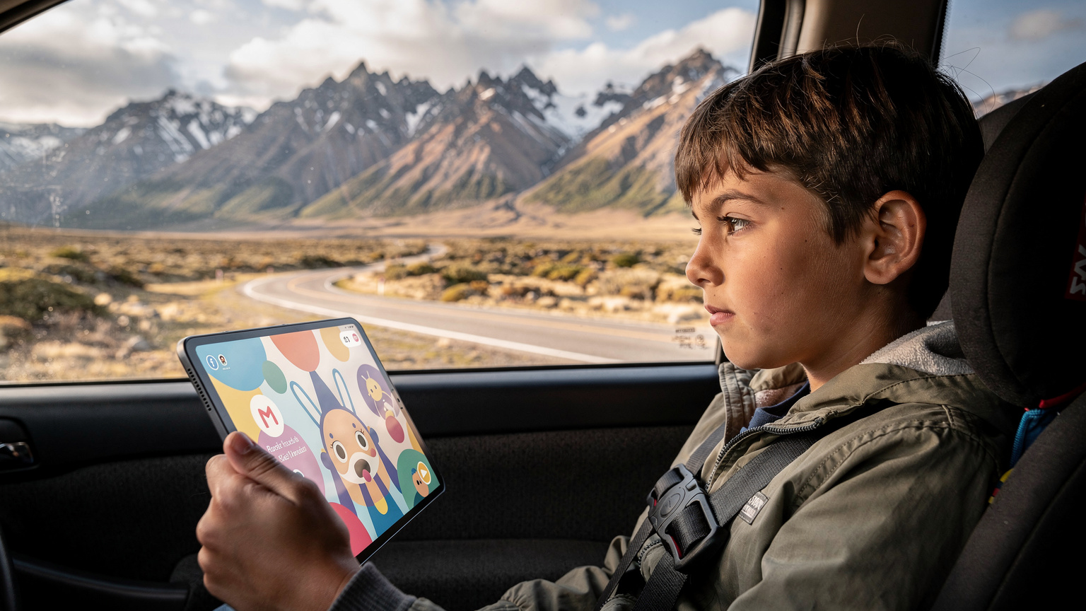 En forældre sidder i en bil i Patagonien og holder en tablet, der viser en streamingtjeneste, mens et barn kigger ud af vinduet, et symbol på underholdning på farten.