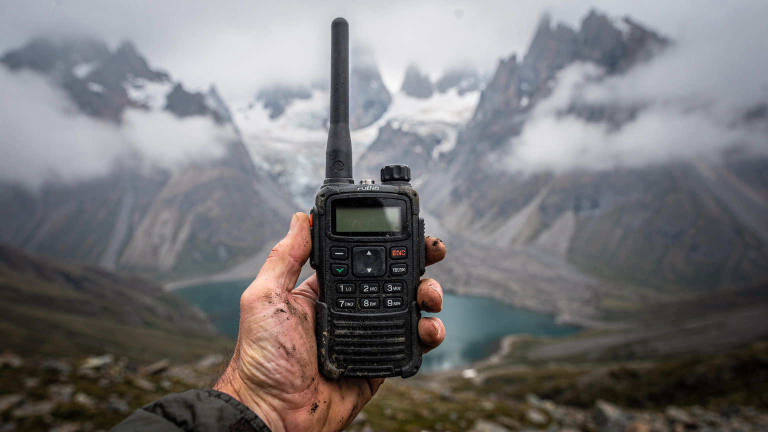 En satellittelefon holdes af en hånd mod en baggrund af tågede bjerge og en fjern sø i Patagonien.