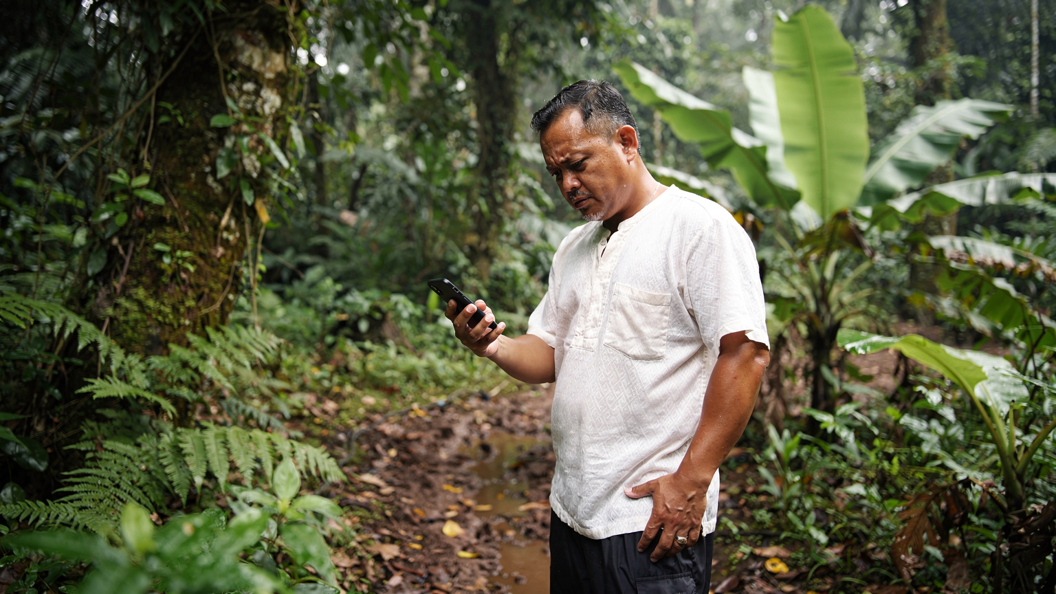 Un guide local thaïlandais en chemise traditionnelle vérifie son téléphone avec une expression préoccupée dans une clairière de la jungle de Chiang Mai, le signal étant faible.