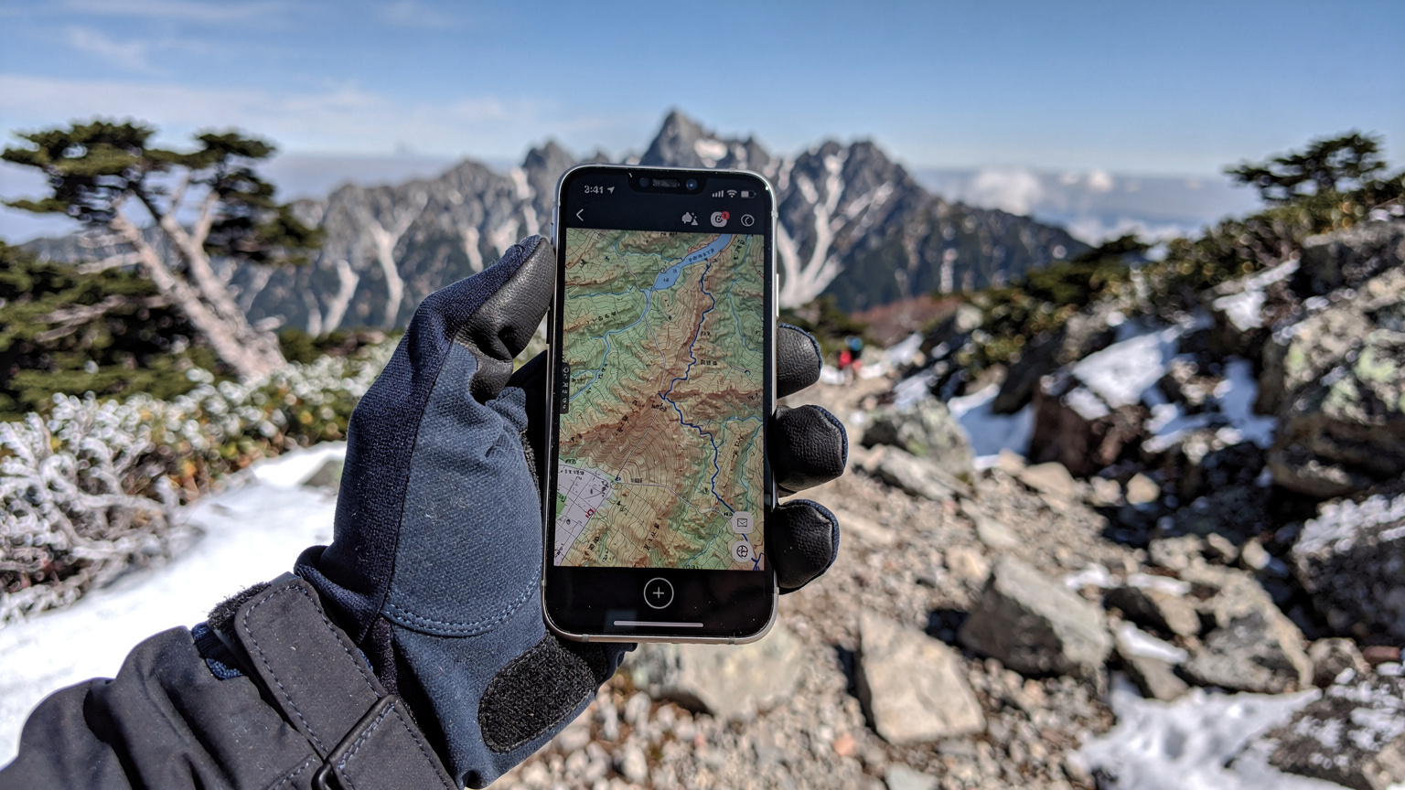 A gloved hand holds a smartphone displaying a hiking map application, with a blurred background of a snow-covered mountain path and sparse pine trees in the Japanese Alps.