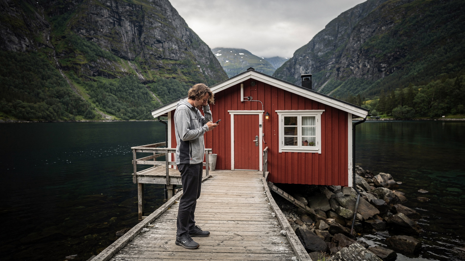 En person forsøker å få mobildekning med en smarttelefon på en trebrygge ved en hytte i en isolert norsk fjord.