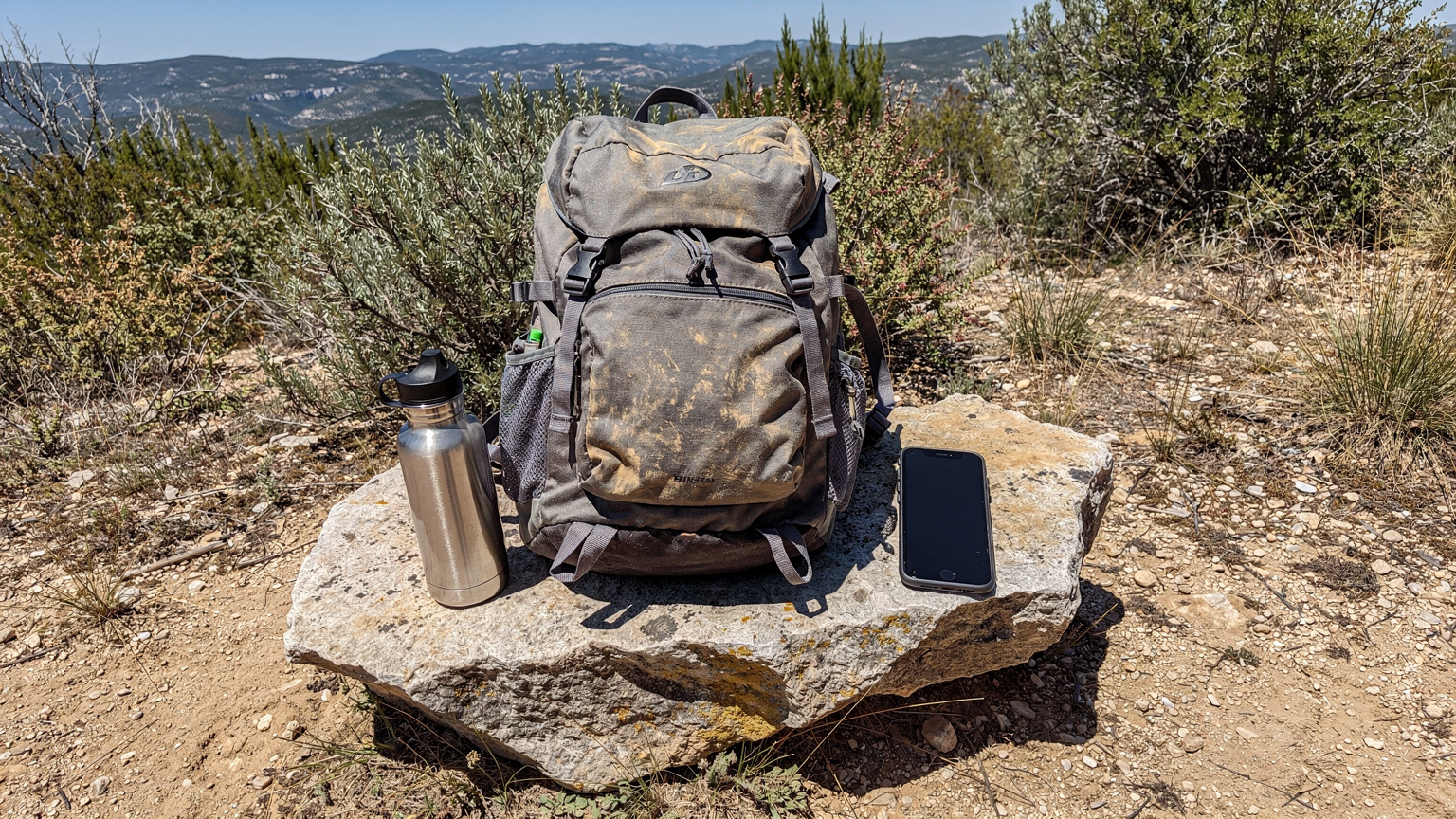 Un sac à dos de randonnée usagé posé sur un rocher, avec une gourde en métal réutilisable et un smartphone à côté, devant un sentier de terre et une vue lointaine sur des collines provençales.