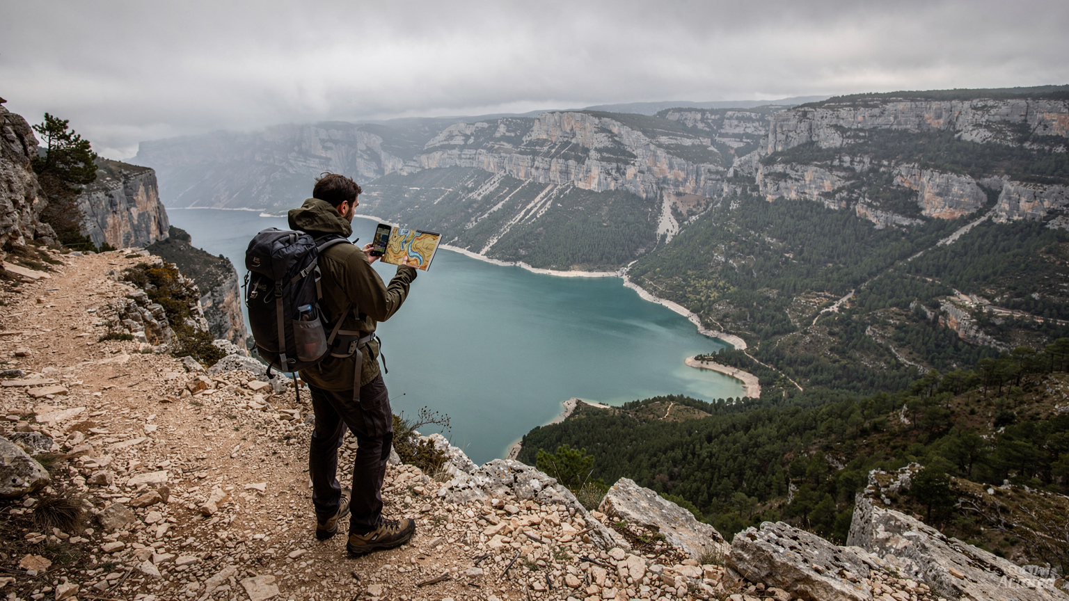 Un randonneur solitaire, vu de dos, consultant son smartphone avec une carte hors ligne affichée, debout sur un sentier rocailleux surplombant un lac turquoise et des forêts denses sous un ciel menaçant.