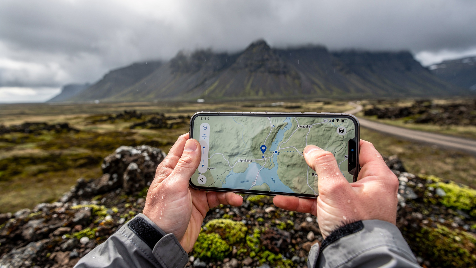 En person som holder en mobiltelefon med et offline-kart vist på skjermen, mens været slår om i det fjerne over et islandsk landskap.