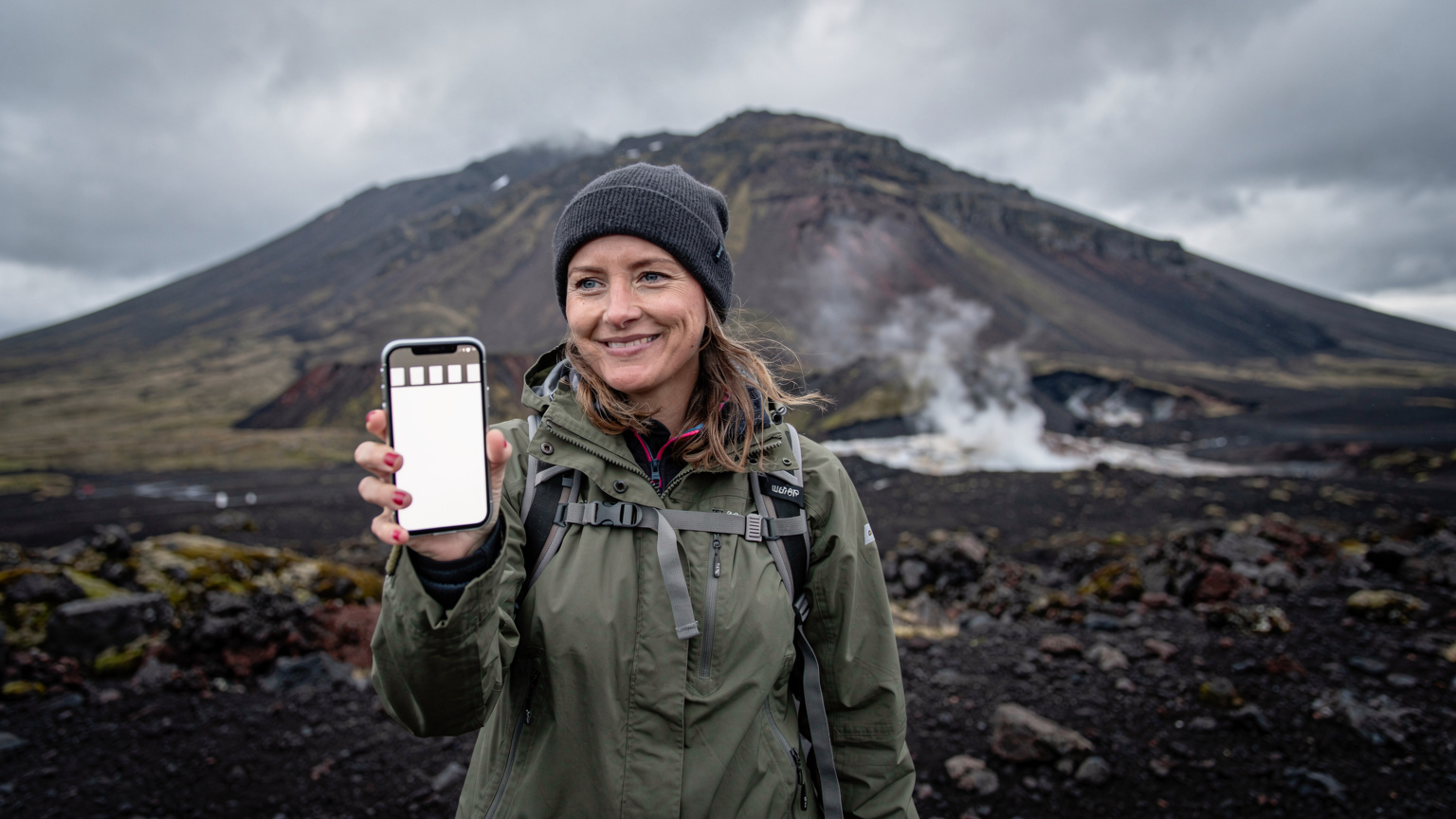 En kvinne i turklær med en mobiltelefon som viser full dekning, stående ved foten av en vulkan på Island.