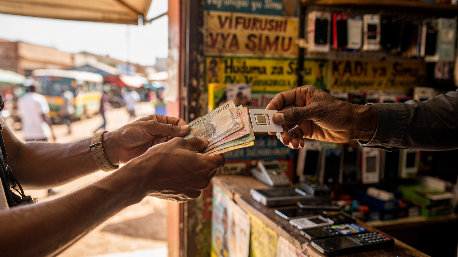 Un primo piano di mani che scambiano una SIM fisica tra un viaggiatore e un commesso locale in un piccolo negozio di telefonia in una strada affollata di Arusha, con insegne swahili e prodotti locali sullo sfondo.