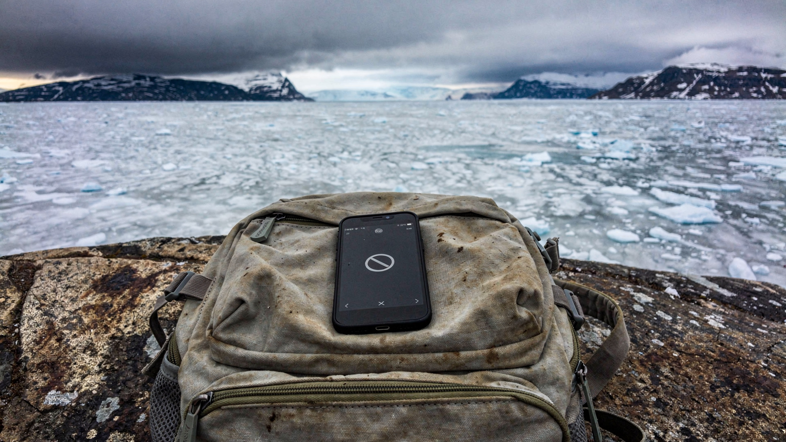 En rygsækspakke med en mobiltelefon, der viser et ingen-signal-ikon, ligger på en klippeflade med en frossen fjord i baggrunden, hvilket illustrerer begrænset dækning i fjerne områder.