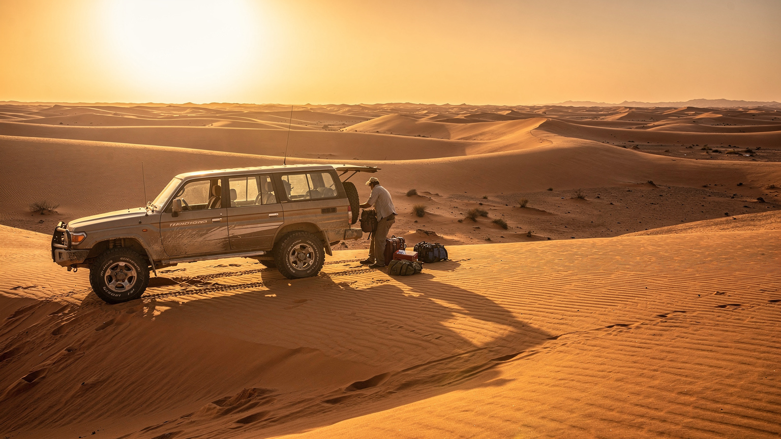 Un fuoristrada 4x4 robusto è parcheggiato su una duna di sabbia, mentre una persona sta scaricando bagagli e attrezzature da campeggio da un lato del veicolo, con il sole all'alba che illumina la scena nel deserto del Sahara.
