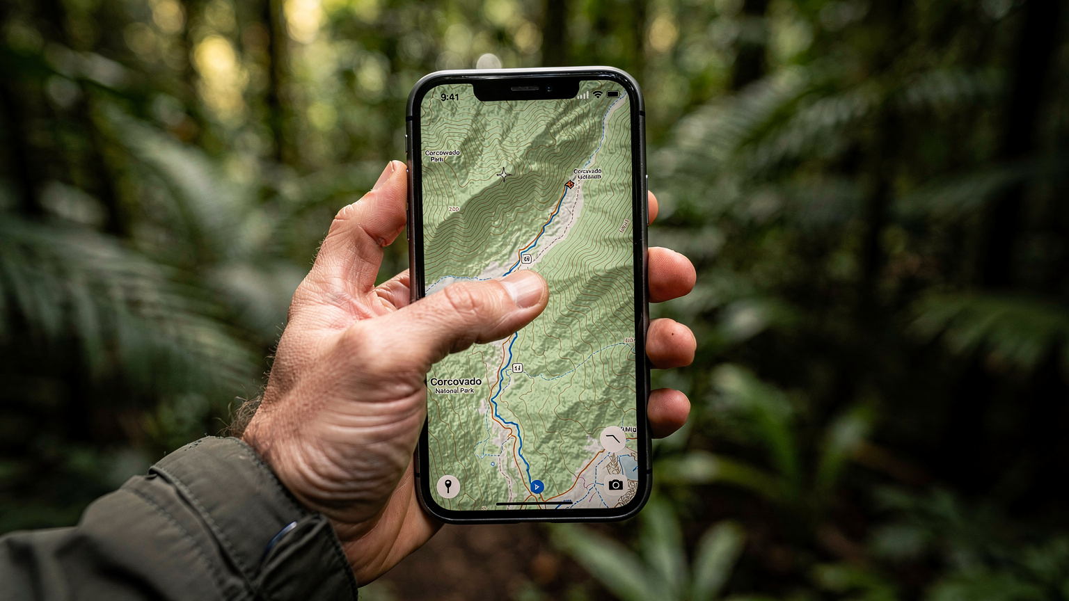 En hand håller en smartphone som visar en nedladdad offlinekarta över Corcovado nationalpark, omgiven av frodig regnskogsvegetation, med fingret som pekar på en markerad stig.