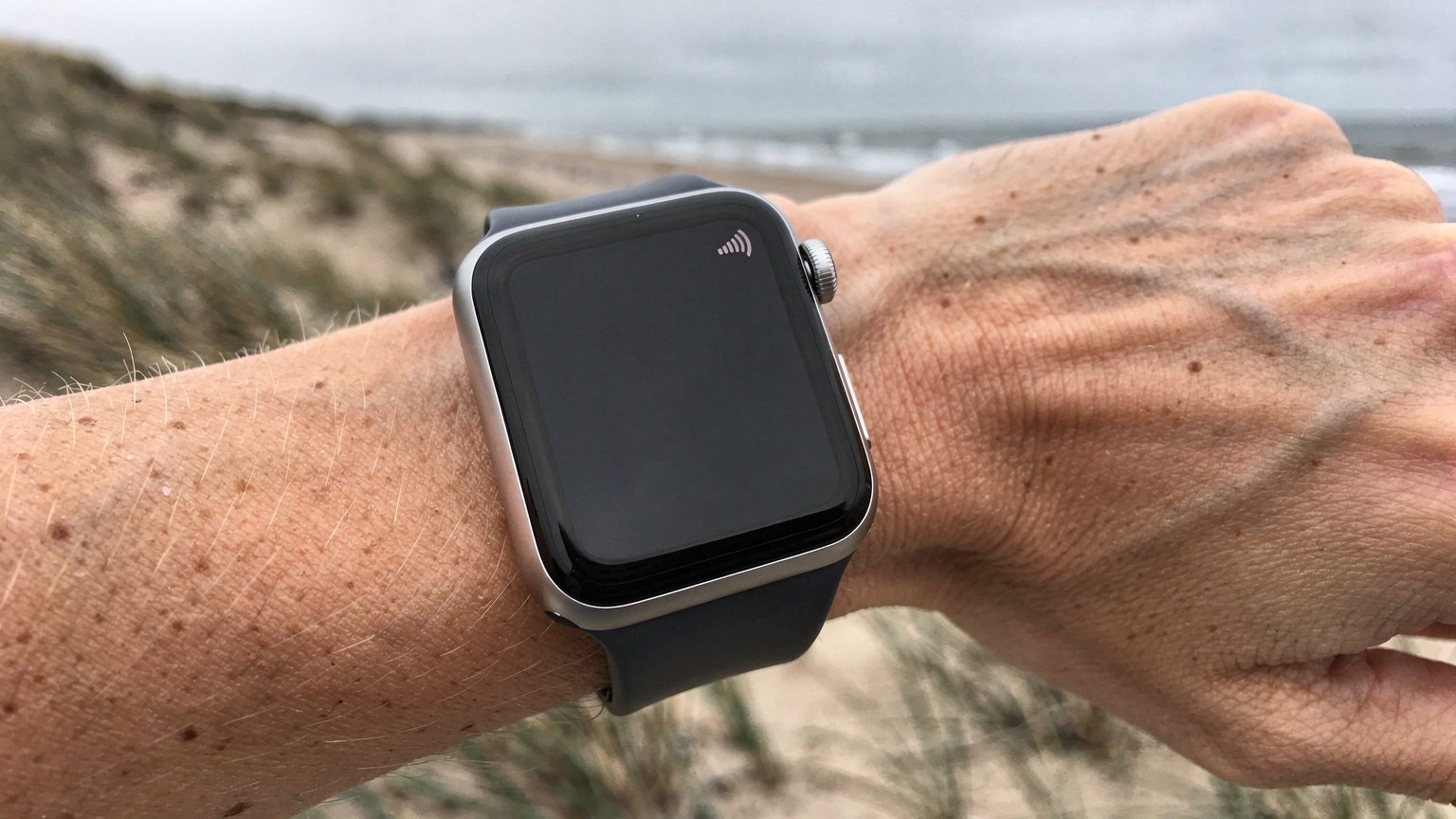 En kvindes hånd med et Apple Watch, der viser svagt signal, mens hun står ved en forblæst Vesterhavskyst med klitter i baggrunden.