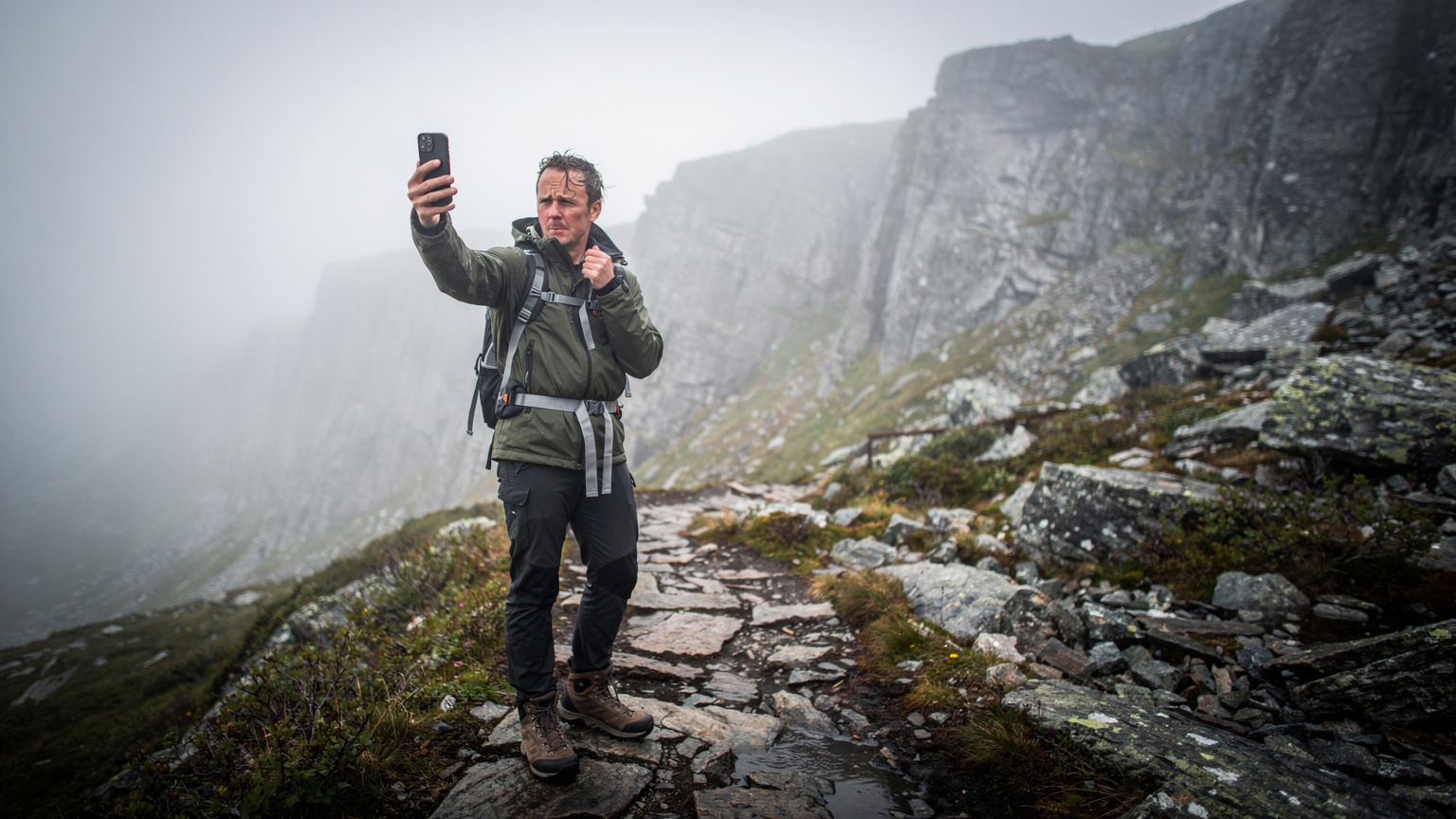 En vandrer tjekker sin telefon for signal, omgivet af tæt tåge og klipper i et fjernt norsk fjeldområde, med en bekymret mine.
