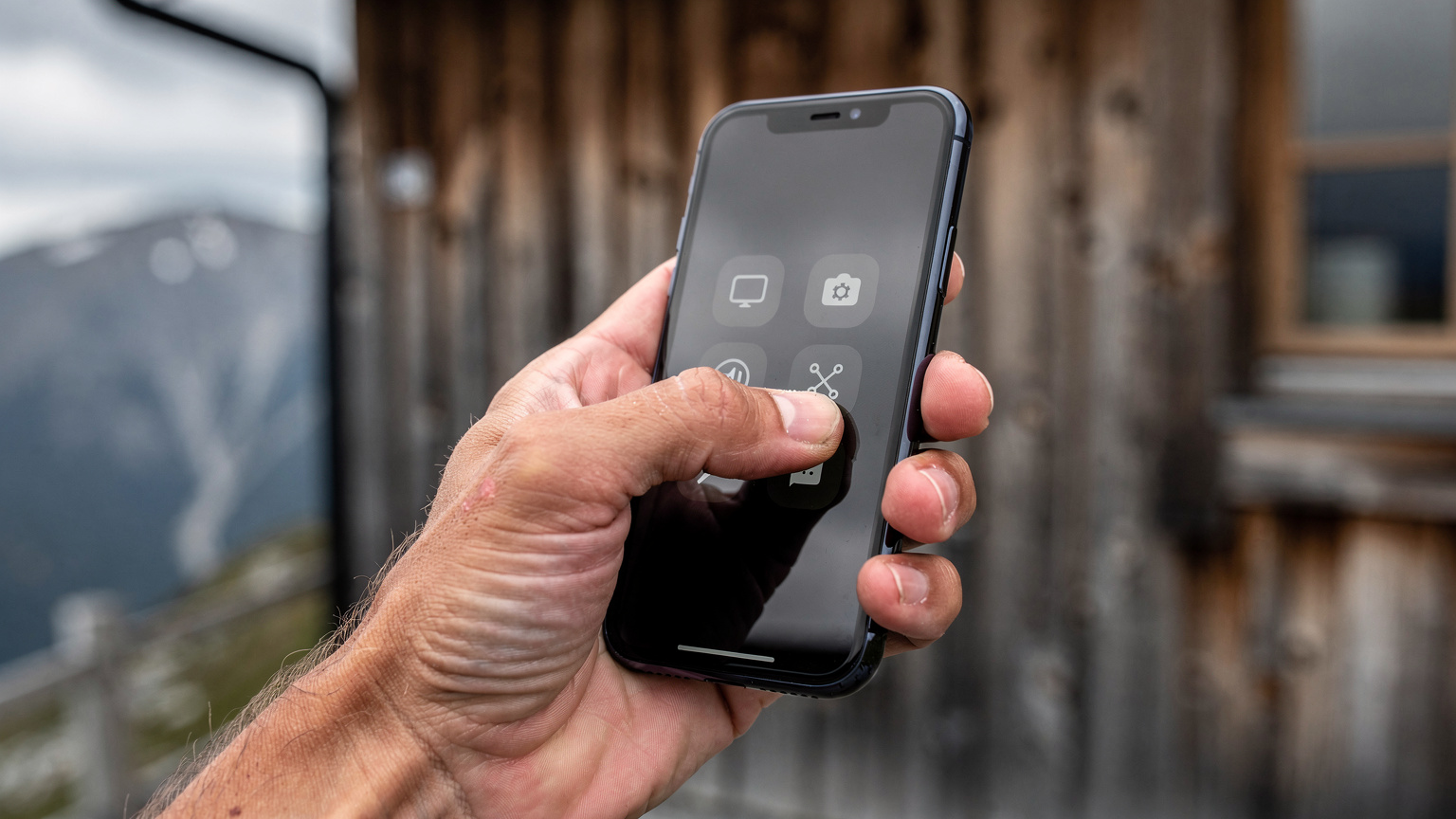Nærbillede af en hånd, der holder en smartphone og trykker på skærmen for at vælge et netværk i telefonens indstillinger, med et sløret baggrund af en træhytte i fjeldet.