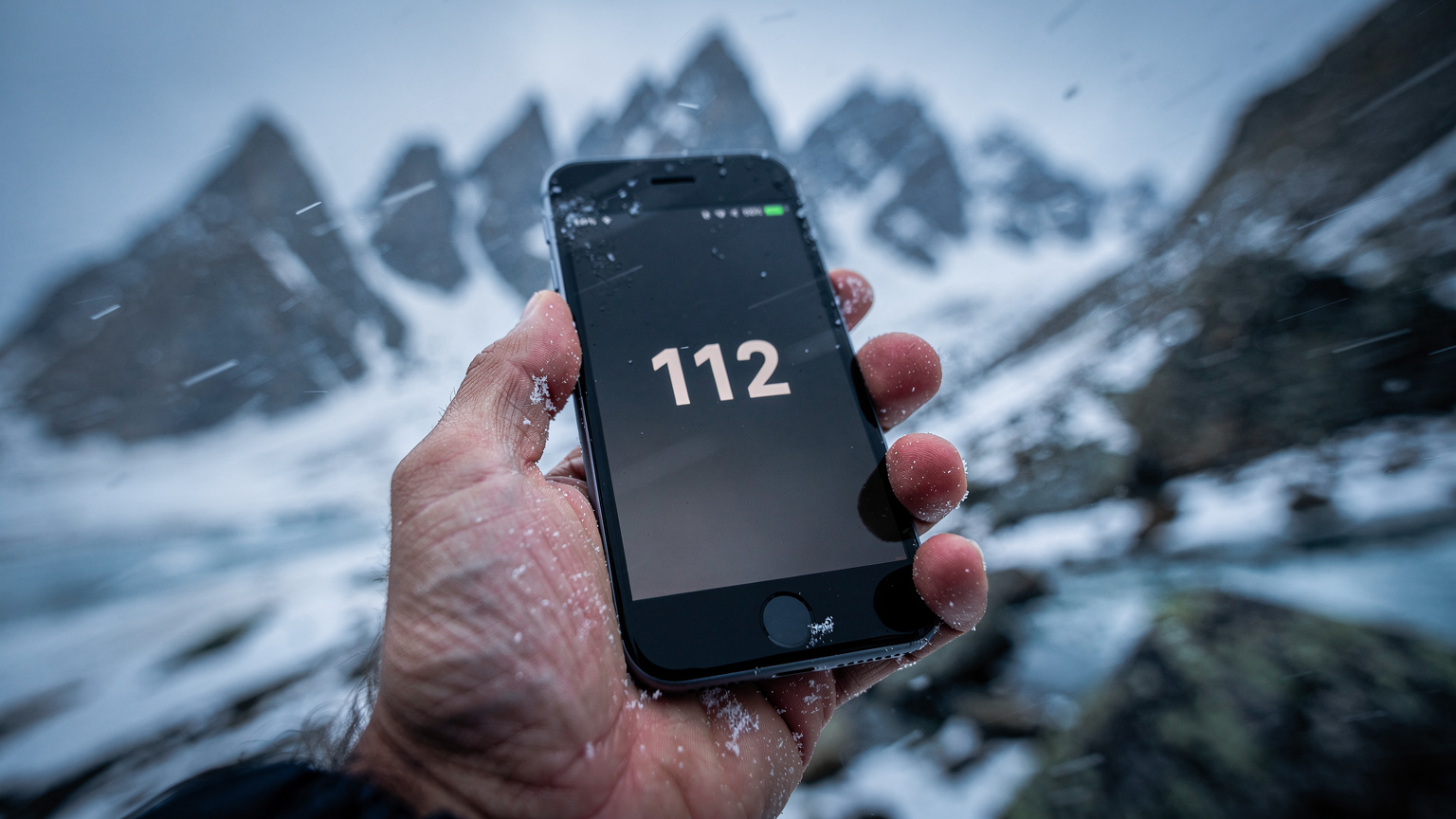 En hånd holder en smartphone med en nødopkaldsskærm vist, mens den ryster let i en kold og blæsende fjeldmiljø med snefnug i luften.