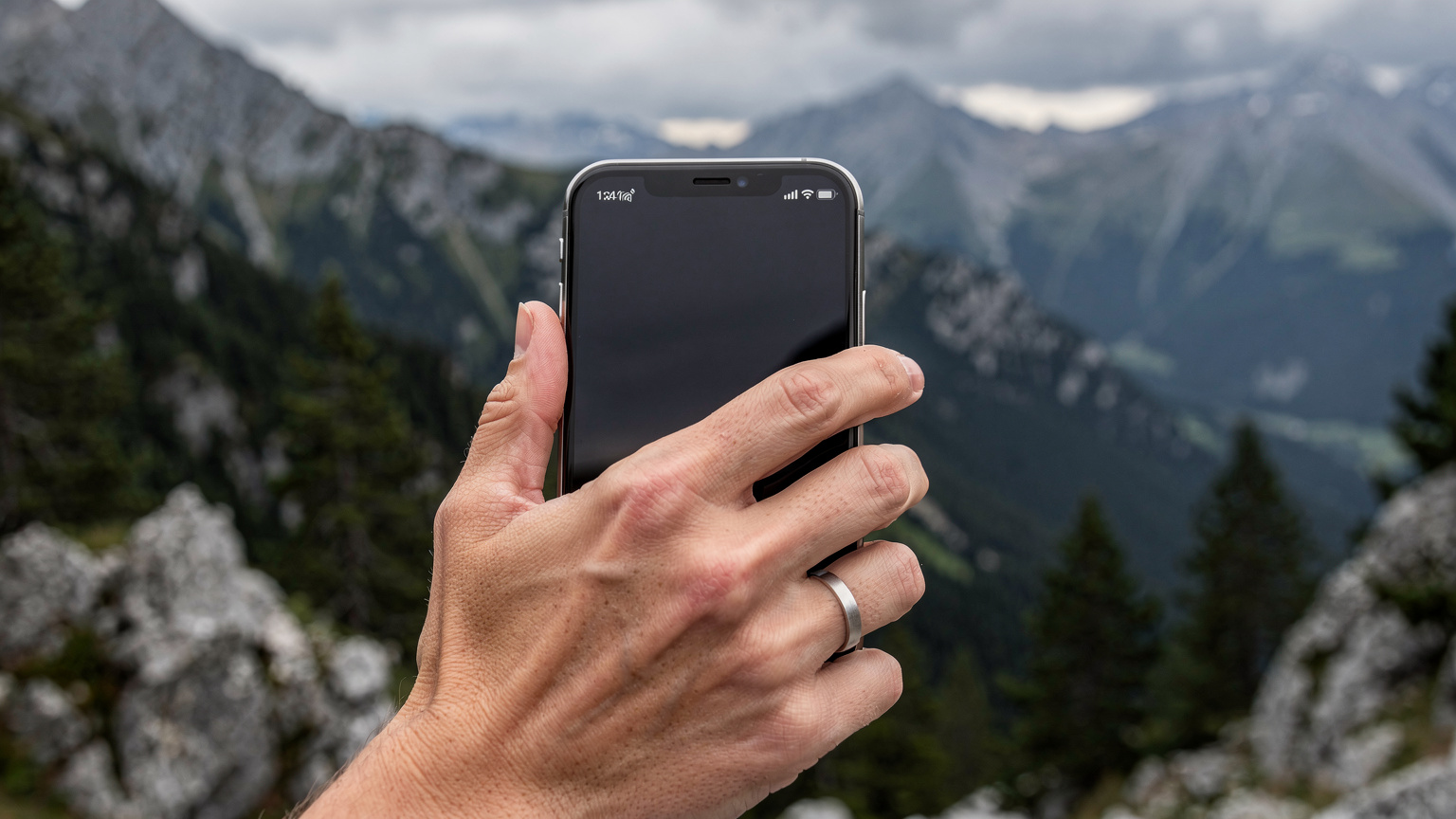 Un smartphone affichant un indicateur de signal faible est tenu par une main d'adulte, avec un paysage de montagne français flou en arrière-plan, suggérant une zone reculée.
